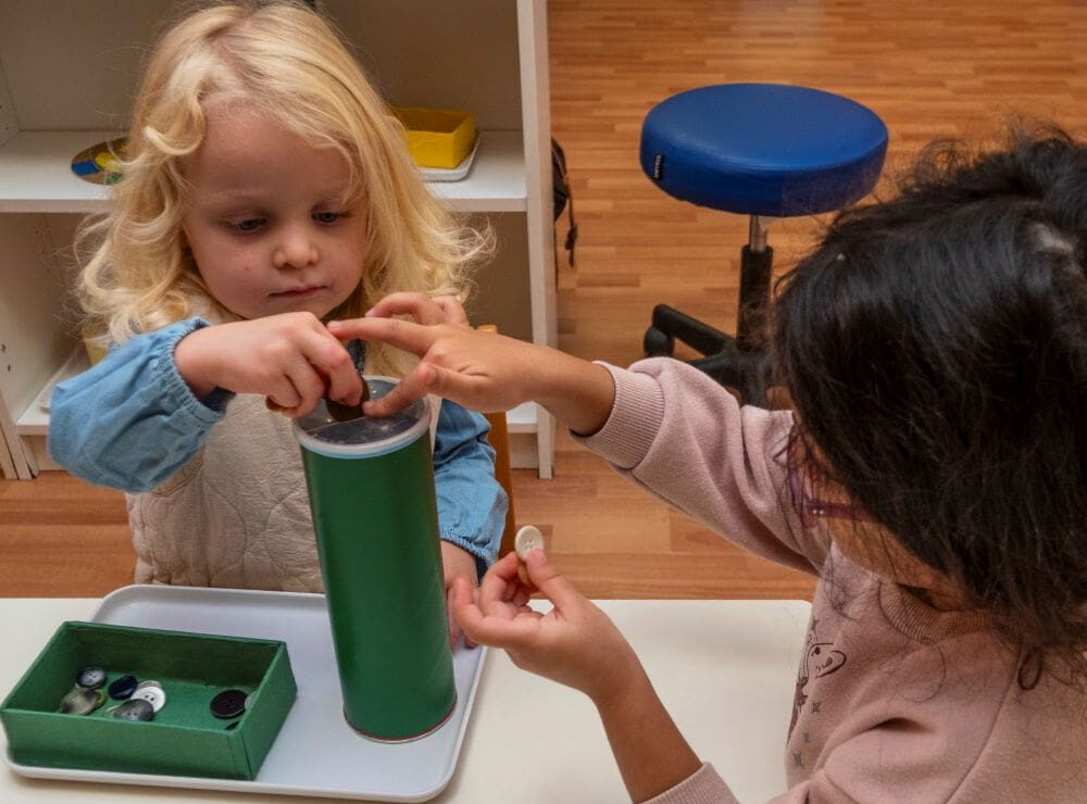 Zwei Kinder schieben verschiedene Knöpfe mit Feingefühl durch einen Schlitz in eine Dose.
