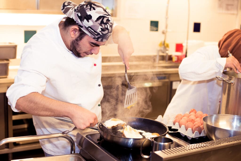 Ein junger Mann brät Spiegeleier in einer großen Pfanne. Er macht eine Kochausbildung in der ITA Wien.