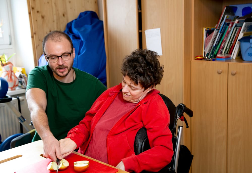 Eine Klientin und ein Mitarbeiter einer vollbetreuten Wohngemeinschaft von KoMiT sitzen gemeinsam am Tisch und schneiden einen Apfel auf.